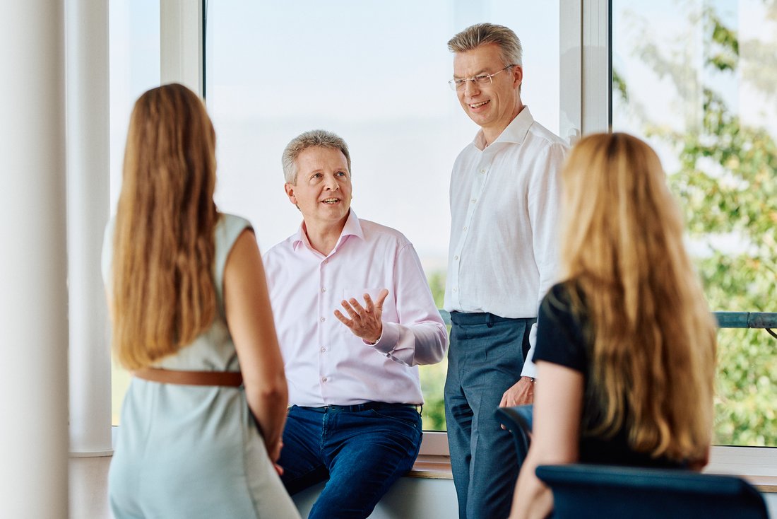 Vier Personen befinden sich in einem Besprechungsraum. Zwei Männer sprechen vor einer großen Fensterfront mit zwei sitzenden Frauen. Die Gruppe wirkt in ein Gespräch oder eine Präsentation vertieft, in einer hellen und modernen Büroumgebung.