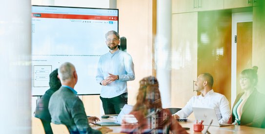 Kolleg:innen in einem modernen Besprechnungsraum. Ein Mitarbeitender präsentiert seine Ergebnisse an einem Bildschirm.