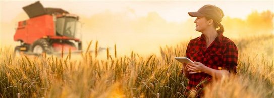 Eine Landwirtin steht mit Tablet in der Hand in einem Feld bei Sonnenschein. Im Hintergrund steht eine landwirtschaftliche Maschine. 